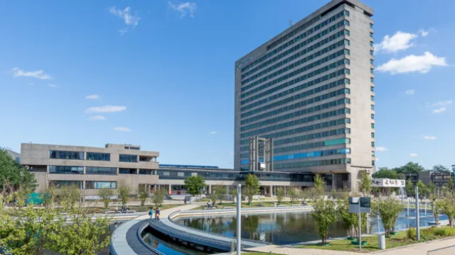 Image of a tall long rectangular building next to a large body of water with walkways and a bright blue sky with some clouds