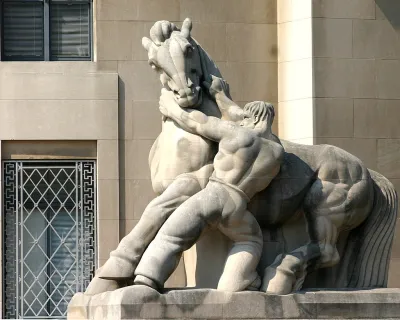 A stone statue of a shirtless muscular man wrestling with a powerfully built horse, his hands on the horse's neck, pulling downward.