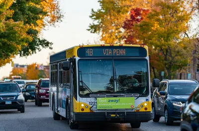 Front of city bus driving down tree-lined street, surrounded by cars.