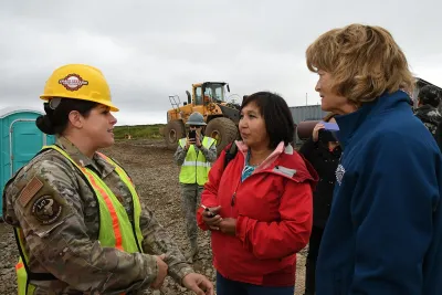Three people, one in fatigues and hard hat, standing in front of bulldozer discussing relocation plans in Newtok, Alaska.