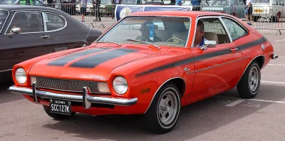 Red 1972 Ford Pinto with black racing stripes on display with man sitting in driver's seat.