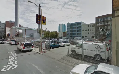 A surface parking lot, full of cars, is located between tall buildings and urban density.