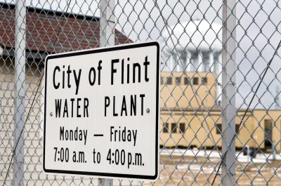 Close-up of sign on chain link fence "City of Flint Water Plant" with hours.