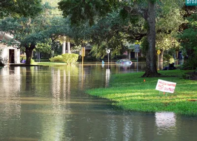 Flooded residential street with houses, yards, and trees on each side, a yard sign that reads "high water, no outlet," and a flooded car in the distance.