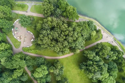 Bird's eye view of park with green trees, concrete walkways, and benches on lake or riverfront.