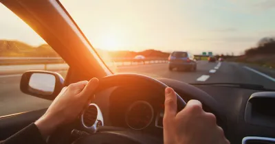 Cose-up of person's hands on steering wheel driving down highway with sunset sky.