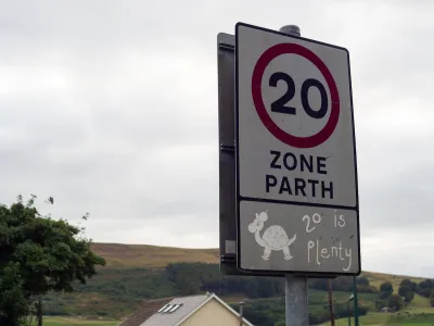 Close-up of white 20 kph speed limit sign in Europe with cartoon turtle and '20 is plenty' sticker underneath.