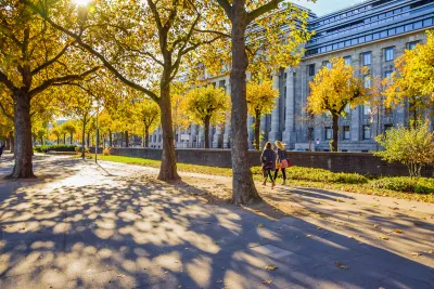 Two women walking on wide walkway among large trees next to neoclassical building in Cologne, Germany.