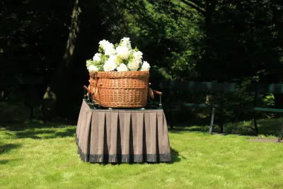 Woven willow casket with white flowers on top sits on table with pleated tablecloth on green lawn