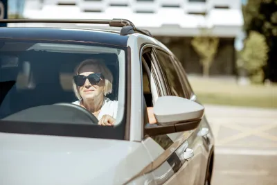 Senior woman wearing sunglasses sitting in the driver's seat of a silver car.