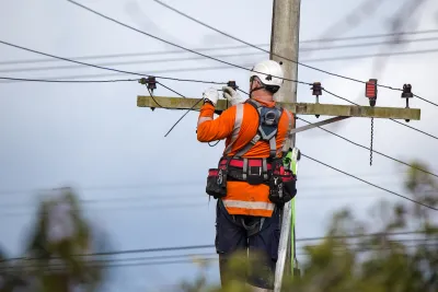 Worker in orange shirt and white helmet works at top of power line.
