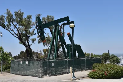 Green oil well pump jack surrounded by fence on a hill overlooking neighborhood.
