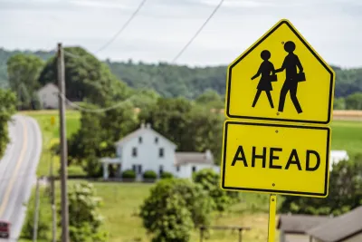 Close-up of yellow and black 'Pedestrians Ahead' sign with blurred farmhouse and road in background.