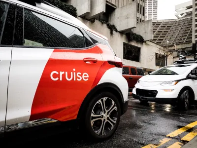 White and red Cruise autonomous car on San Francisco street with concrete Brutalist-style building in background on grey, rainy day.