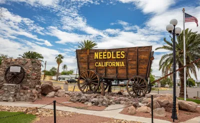 Sign for Needles, California installed on wooden wagon with residential neighborhood visible in background.