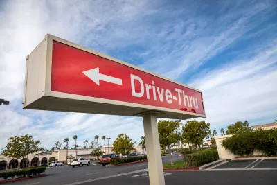 Close-up of red sign with white "Drive-Thru" text and arrow pointing left against partly cloudy blue sky.