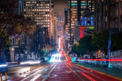 Blurred lights of traffic in motion on San Francisco street with high-rise buildings and Bay Bridge visible in background at night.
