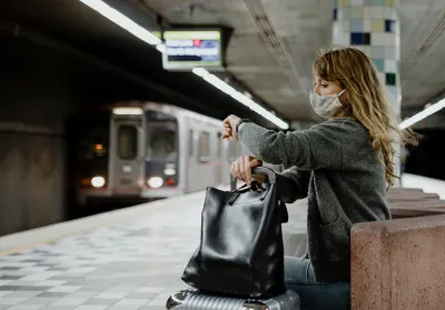 Woman with long hair wearing Covid mask sitting on underground train station bench looking at her watch as subway train approaches in background at Hollywood/Western station in Los Angeles, California.