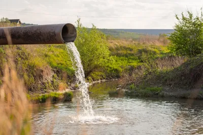 Large metal pipe discharging water or wastewater into creek or body of water with shrubs and field in background.
