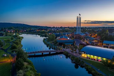 Aerial view of Bend, Oregon with river in foreground and factory smokestacks with American flags on top against sunset sky.