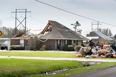 Exterior side view of a single-story brick home missing back half of roof and a debris pile in the front yard.