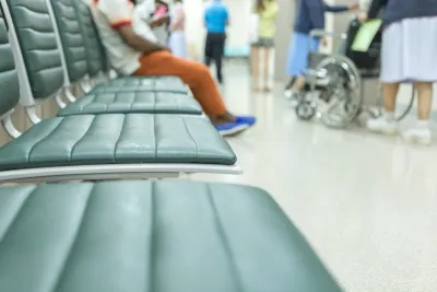 Close-up of blurred empty vinyl chairs with person sitting in background and wheelchair visible in hospital waiting room.