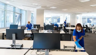 Professional cleaners wearing medical masks and gloves cleaning desks in large empty office.