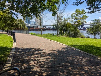 Brick path through an urban park shaded by trees with a river on the righhand side and high-rise buildings in the distance.