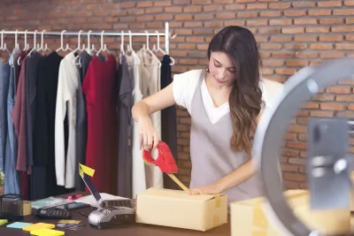 Woman taping up a brown paper package with rack of clothing in background.