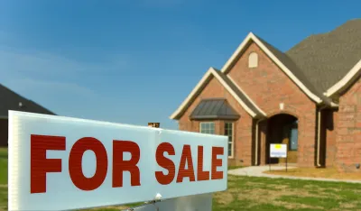 Close-up of white sign with red FOR SALE letters and blurred brick single-family home in background.