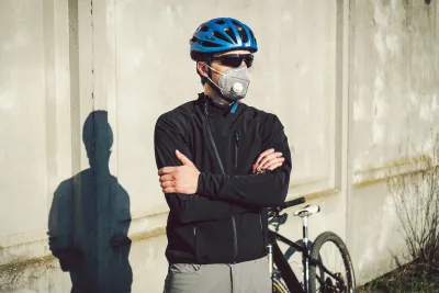 Male cyclists wearing helmet, sunglasses, and N95 mask stands with arms crossed in front of his bike