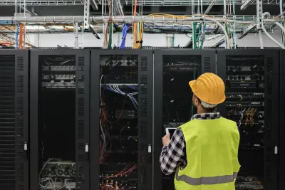Worker in yellow safety vest and hard hat looks up at servers in data center.