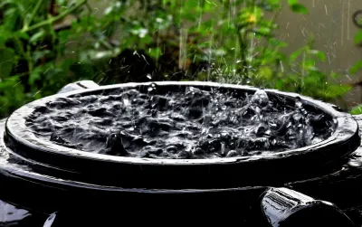 Close-up of top of black rain barrel full of water with raindrops falling on surface.