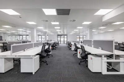 Large empty open-plan office with rows of white desks and black chairs under bright fluorescent lighting
