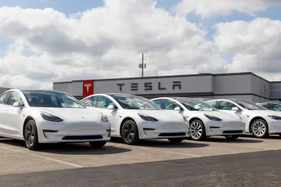 Row of white Tesla electric cars in parking lot with dealership building with Tesla logo in background.