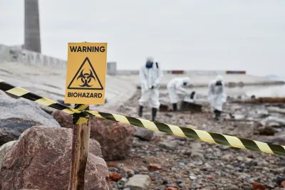 Close-up of yellow "Warning Biohazard" sign with people in hazmat suits cleaning up in background.