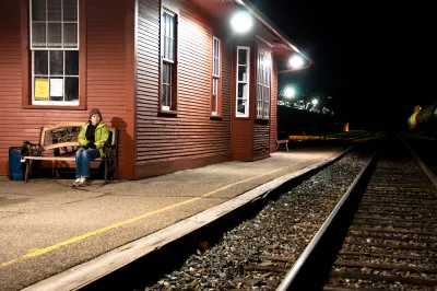 Womanin yellow jacket sitting on bench at outdoor train station with red wood depot building at night.
