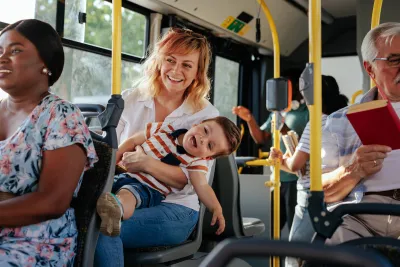 Woma holding small boy on lap sitting on public bus with many other passengers around them.