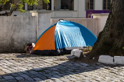 A orange-and-blue tent set up next to a tree in a paved courtyard and a cardboard box of posessions next to the tent entrance.
