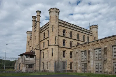Deralict stone prison building with turrets and a chain link fence in foreground, under a gray, cloudy sky.