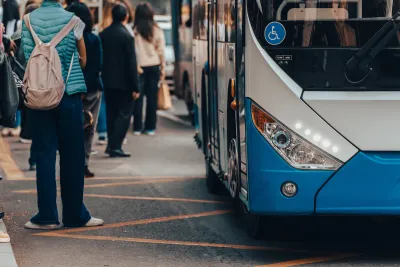Close-up of front side of blue and white bus with accessibility sticker and people wearing backpacks waiting outside of bus.