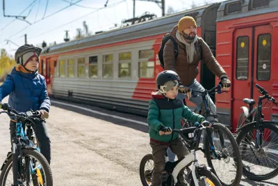 Man and two children on bikes next to red and silver train on sunny day.