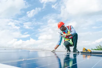 Worker wearing orange hard hat and vest over light blue long-sleeved shirt and jeans inspects rooftop solar panel