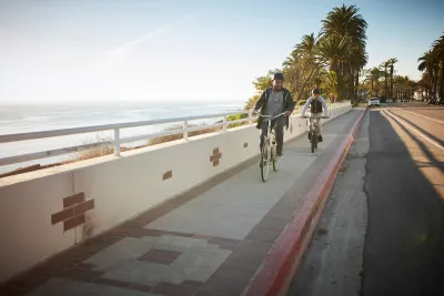 Father and son riding bikes on a sidewalk next to a road withough bike lane on California coast with palm trees in background at golden hour.
