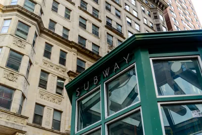Green subway station entrance kiosk with brick buildings in background.