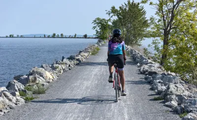 View from behind of woman wearing helmet on bicycle riding on concrete causeway with water on either side.