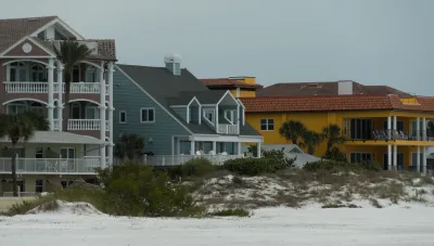 Three colorful, large beachfront homes, one khaki, one blue, and one yellow, with a small dune in front and flat sand in foreground.