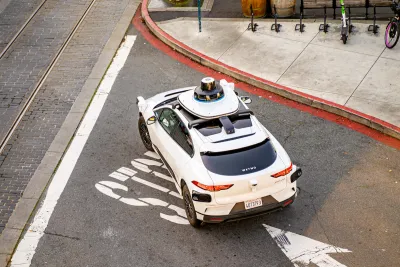 Overhead view of white Waymo self-driving car in San Francisco, California with shared bikes and scooters visible on adjacent sidewalk.