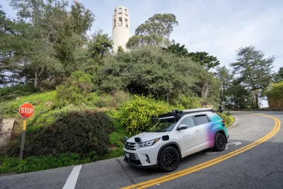White self-driving car at stop sign coming down hill with Coit Tower in background in San Francisco, California.