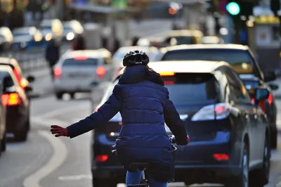 Rear view of woman in blue coat riding bike on busy road signals with left hand to merge into lane of traffic.